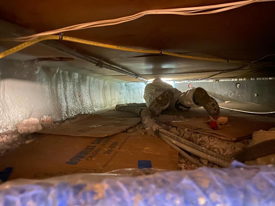 A technician in protective gear crawling on cardboard to move forward on a ceiling being insulated.