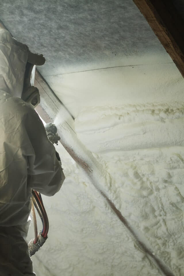 A man in protective gear applying spray foam insulation to the walls of an attic.